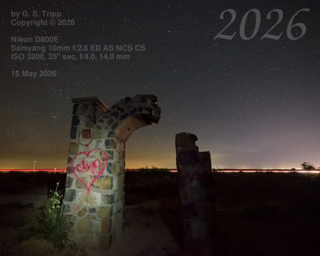 A long-exposure night photograph of a ruined stone archway and pillar under a clear, star-filled sky. The stone pillar in the foreground features pink heart-shaped graffiti. In the distance, a horizontal streak of red and white light from a highway glows against the dark horizon. Technical camera settings and the year 2026 are overlaid in text on the corners of the image.
